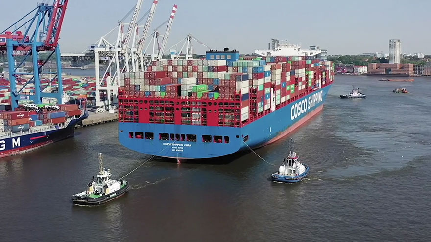 Mit Containern beladenes Schiff, davor zwei Schlepper im Hafen.